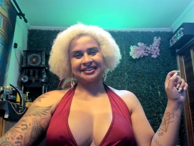 MarianaJade, curvy with large breasts, wearing a deep red halter top, has platinum blonde curly hair, tattooed arms, and a nose ring. Smiling, holding a cigarette, in a dimly lit room.