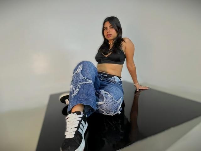 Celeste Morel, una donna dai capelli scuri, seduta su una superficie nera, con le gambe divaricate, indossa jeans blu strappati e un reggiseno sportivo nero. Sneakers Adidas bianche, che guardano verso la fotocamera, tensione sessuale, posa sicura, stile casual..