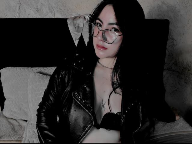 JeniferPowell, à la peau pâle et aux cheveux noirs, portant des lunettes et une veste en cuir noir, avec un soutien-gorge noir visible, allongée sur un lit avec des oreillers gris, un regard séduisant, des lèvres légèrement entrouvertes, des ombres sombres..