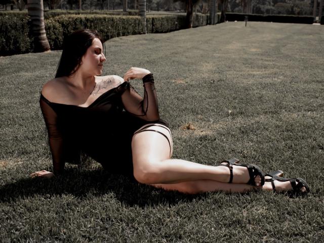 Jenifer Powell est allongée sur l'herbe, les jambes écartées, portant un haut noir transparent et une culotte noire, exposant sa peau pâle, ainsi que des talons noirs. Tatouages sur le bras droit, arrière-plan ombragé, forte tension sexuelle, pose séduisante..