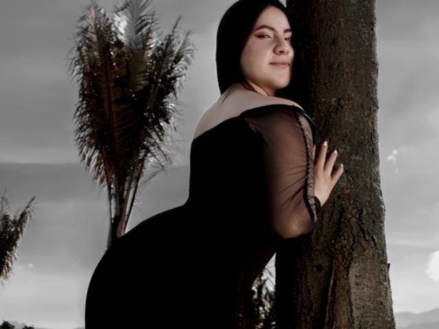 Photographie de Jenifer Powell appuyée contre un arbre, portant une robe noire transparente avec un col blanc, et regardant au-dessus de son épaule. épaule. L'arrière-plan comprend de grands arbres et un ciel nuageux. Femme séduisante, aux cheveux foncés..