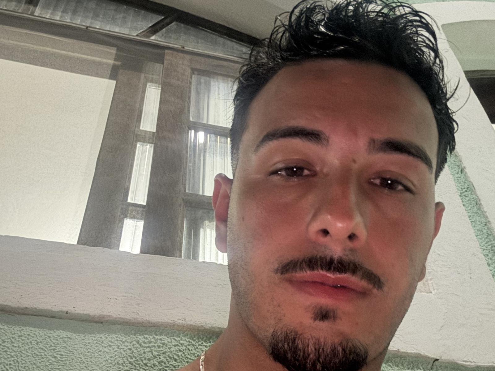 Close-up de Crispin69 com o rosto com barba rala, cabelo escuro e desalinhado, e um leve sorriso. Ele está vestindo uma camisa branca, em pé perto de grandes janelas, com uma parede verde ao fundo. Muito atraente..