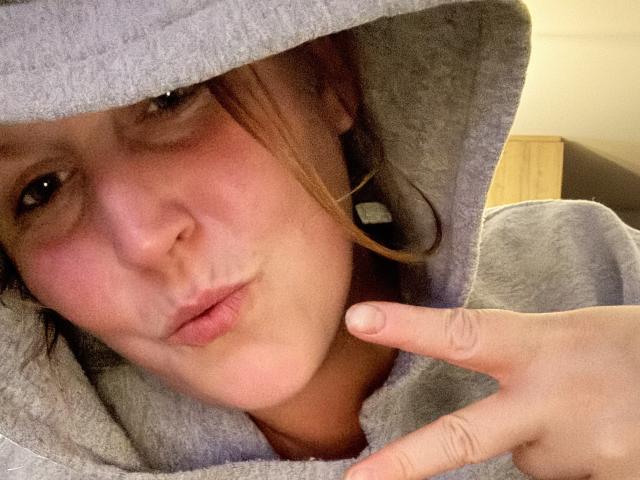 Close-up photo of Yssa69, face partially hidden under a gray hoodie, right hand near face with index finger raised, slightly parted lips, reddish-brown hair, soft light, casual expression.