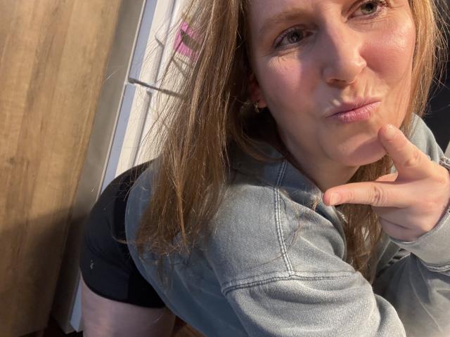 Primo piano di Yssa69 con lunghi capelli castani, pelle chiara e occhi azzurri, che indossa una giacca di jeans e guarda direttamente verso... fotografia di una donna con un dito vicino alla bocca, la luce del sole che illumina il suo viso.