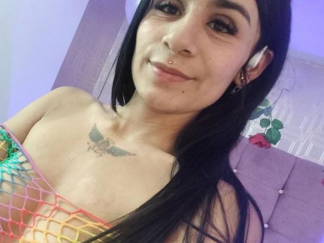 Photo en gros plan d'AmberLuxefg, souriante, avec des cheveux foncés, un piercing au nez, un haut en résille coloré et un petit tatouage sur la poitrine. Peau claire, écouteurs dans les oreilles, tension sexuelle subtile, pose décontractée, expression détendue..
