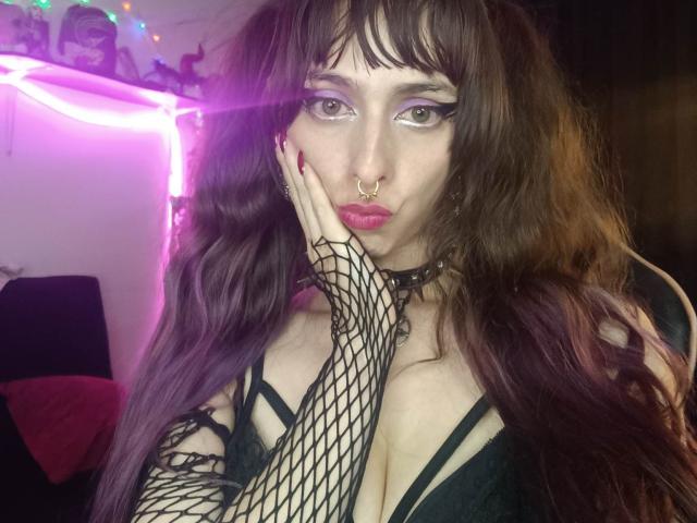 Foto em close-up de KizzuKyuwu, com pele clara, cabelo ondulado escuro e maquiagem carregada. Ela está usando uma luva de rede preta, um colar e uma blusa preta. Olhando sedutoramente, com a mão no rosto, luzes de neon ao fundo..