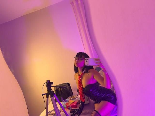 VannesaSaenz with black lace lingerie, red striped tie, and fishnet stockings, kneeling in front of a laptop and webcam, illuminated by pink and purple lights, creating a sexual tension vibe.