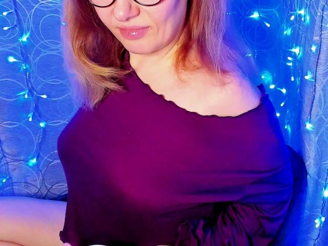 Fotografia de PaulinaSunny com cabelo castanho claro, vestindo uma blusa preta com um ombro descoberto, óculos e luzes azuis. contexto. Seus lábios se abriram ligeiramente, e as luzes azuis criaram uma atmosfera de tensão sexual. Cores vibrantes, pose casual.