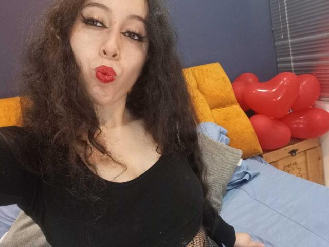 KendraSimons, a pale-skinned woman with long curly black hair, pouts with red lips, wearing a black top, sitting on a bed with yellow pillows and red heart-shaped balloons. Fucking sexy.