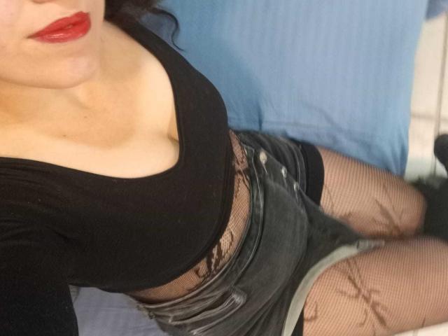 Close-up photo of KendraSimons lying on a blue surface, wearing a black top with a deep neckline, red lipstick, black mesh shorts, and fishnet stockings. Visible cleavage, black leather belt.