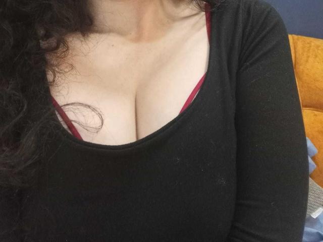 Close-up of KendraSimons' chest, showing large cleavage in a black top with red trim. Black curly hair partially visible. Background includes orange cushion and blue wall. Fucking sexy.