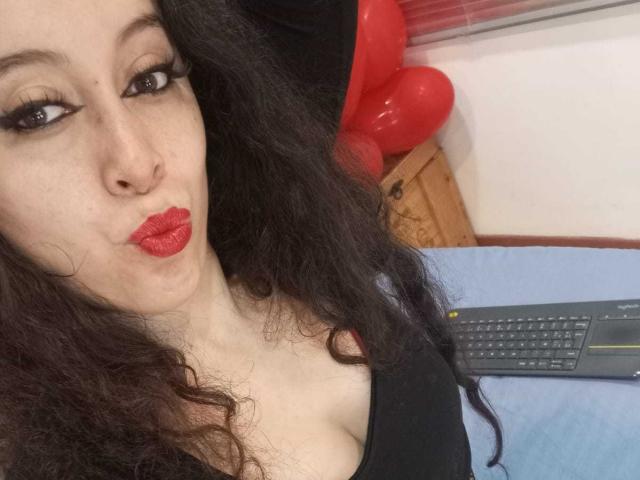 Close-up photo of KendraSimons with long black curly hair, pale skin, and red lipstick, pouting her lips. She has dark eye makeup and wears a black top. Red balloons in the background.
