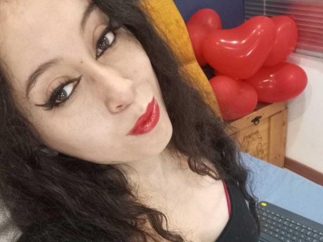 Close-up of KendraSimons with pale skin, black curly hair, and red lipstick, looking seductively at the camera. Background includes red balloon dildos and a wooden box. Fucking sexy vibe.