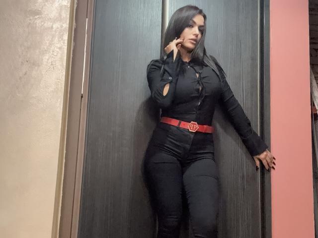 PeachMuse steht vor einer dunklen Holztür, sie trägt ein eng anliegendes schwarzes Hemd und eine Hose mit einem roten Gürtel. Sie berührt ihr Gesicht mit einem sinnlichen Ausdruck. Verdammt heiß. Langes, schwarzes Haar.