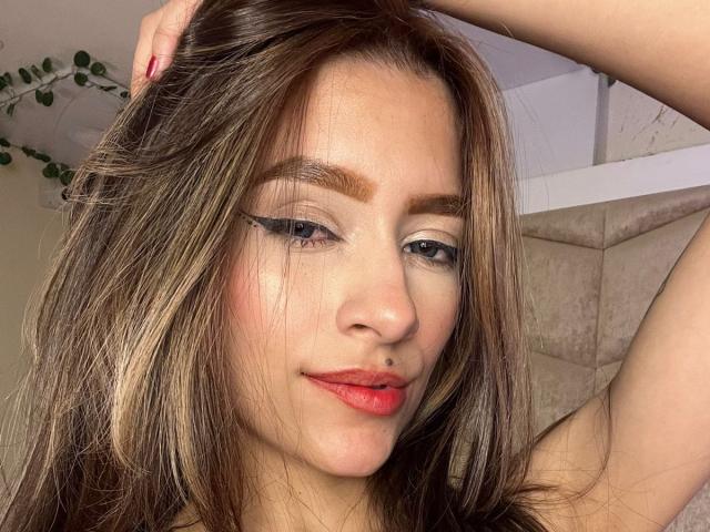 Close-up photo of AfroditaWild, a sexy woman with long brown hair, red lipstick, and blue eye makeup, looking seductively at the camera with her right arm raised. Background includes plant vines.