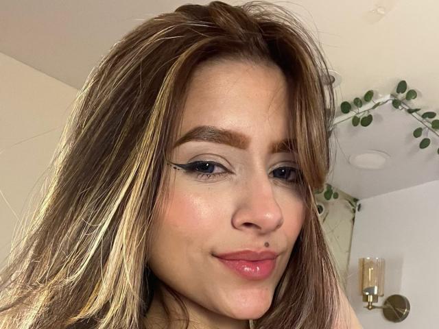 Close-up photo of AfroditaWild, a brunette woman with long, straight hair, black eyeliner, pink lipstick, and a nose piercing. She's smirking slightly, with soft light highlighting her face. Background includes a white wall and a hanging plant.