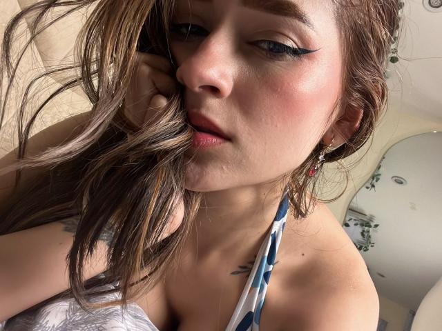 Close-up photo of AfroditaWild, a white woman with long brown hair, heavy black eyeliner, and a white halter top, her face partially shadowed, showing slight cleavage and a serious expression.