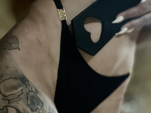 Close-up of DaneeSoHot's side, wearing black lingerie with a cutout. Visible tattoo on lower back, gold clasp on strap, holding black object with white-painted nails. Sexual tension.