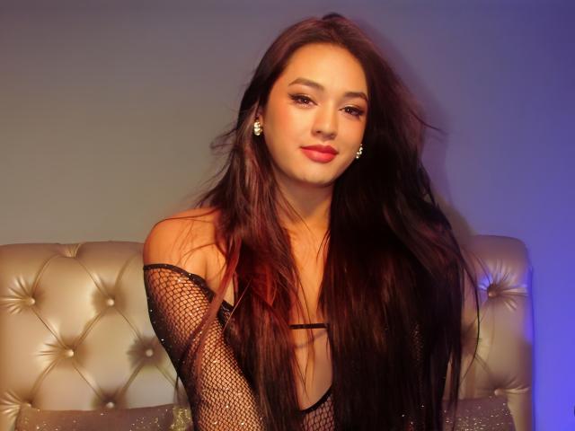 Yessica Belltran, uma mulher atraente com longos cabelos pretos, vestindo uma blusa de rede preta, sentada em um sofá bege com detalhes, azul. luz no lado direito, batom vermelho, expressão confiante, tensão sexual sutil.