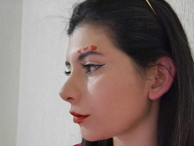 Fotografía de primer plano de JeneKurz, con piel clara, cabello oscuro y marcas rojas en el rostro. El perfil de JeneKurz muestra una mirada intensa, con labios rojos, delineador negro y un pasador dorado. Tensión sexual evidente..