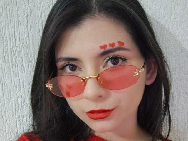 JeneKurz tiene el cabello largo y negro, lápiz labial rojo y tatuajes en forma de corazón rojos sobre las cejas. Ella usa gafas de sol tipo aviador con un tono rojo y monturas doradas. Un labial rojo, increíblemente sexy, que combina con corazones y gafas..