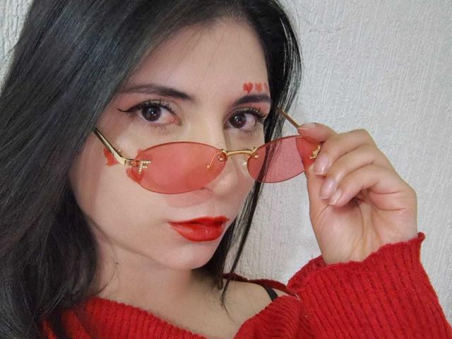 Close-up photo of JeneKurz, wearing a red sweater, holding pink aviator sunglasses, with red lipstick and facial paint dots. She has dark hair, light skin, and a seductive expression.