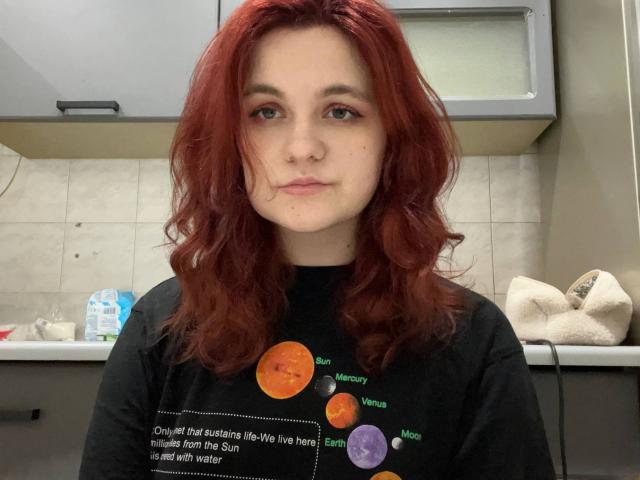 LahomaLoven, con i capelli rossi che le arrivano alle spalle, guarda direttamente l'obiettivo. Indossa una maglietta nera con motivi circolari colorati. Lo sfondo include una cucina con armadietti e sacchetti. Un'espressione molto seria..
