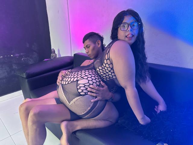 VanesssaXJean with thick, curvy body and large breasts in a fishnet dress, sitting on a muscular guy's lap. She's looking back with glasses, blue light highlighting her face. Sexual tension.