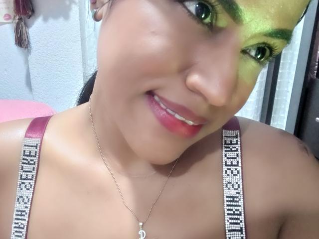 Autoritratto ravvicinato di IvannaDuran, che mostra un viso sorridente con ombretto verde, rossetto rosa e una collana d'argento con ciondolo a forma di "D". Indossando un reggiseno con le spalline con la scritta "CHAMPION". Illuminazione intensa..