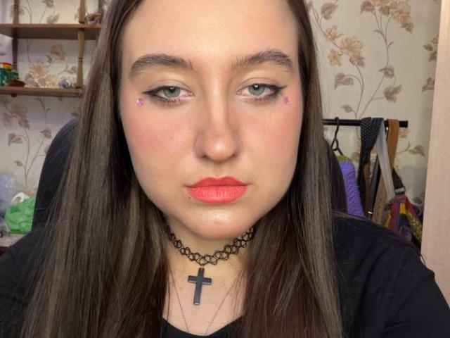 Close-up van SabrineLookyCokky, een vrouw met bleke huid, lang zwart haar, rode lippenstift en een zwarte kruisbedel. Kijkend recht in de camera met een lichte grijns, bloemen behang op de achtergrond..