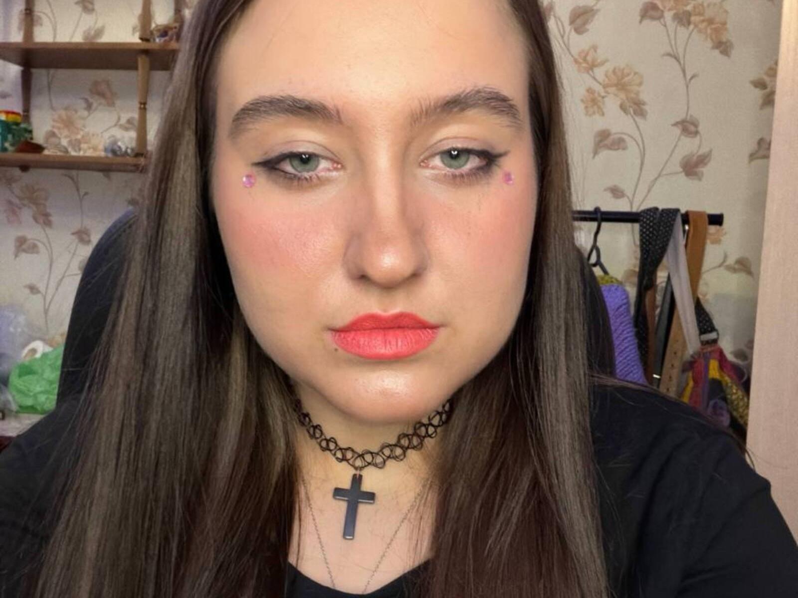 Primer plano de SabrineLookyCokky, una mujer de piel clara con cabello largo y negro, lápiz labial rojo y un collar con una cruz negra. Mirando directamente a la cámara con una ligera sonrisa, papel tapiz floral en el fondo..