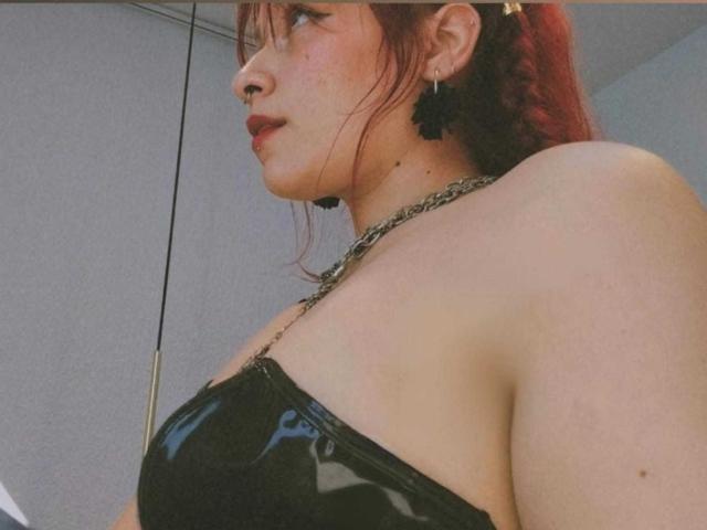 Gros plan de LolyYunno, montrant le profil, avec des cheveux roux tressés, portant un haut en latex noir, un collier en chaîne et des boucles d'oreilles. Peau pâle, sexy, légère rougeur, expression confiante, suggestion de décolleté, tension sexuelle..