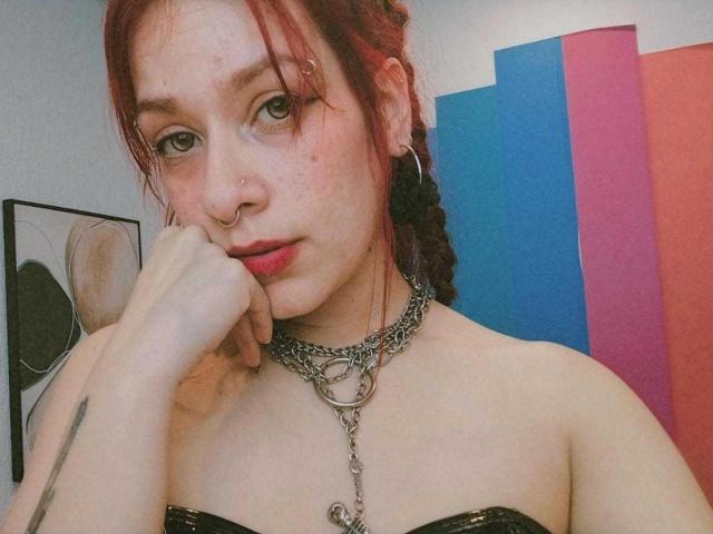 Photographie de LolyYunno avec des cheveux roux, une peau pâle et des taches de rousseur, portant un haut noir sans bretelles, un collier en argent et piercing au nez.
lolyYunno se touche le menton, regardant directement la caméra avec un regard séduisant..