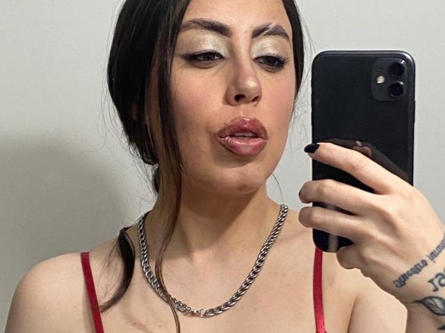 KimyR prend un selfie, tenant un téléphone noir dans sa main droite. Elle a les cheveux foncés, un trait d'eye-liner prononcé et des lèvres rouges brillantes. Elle porte un haut à bretelles rouges et un collier en argent. Elle a un tatouage au poignet gauche. Putain, c'est sexy..