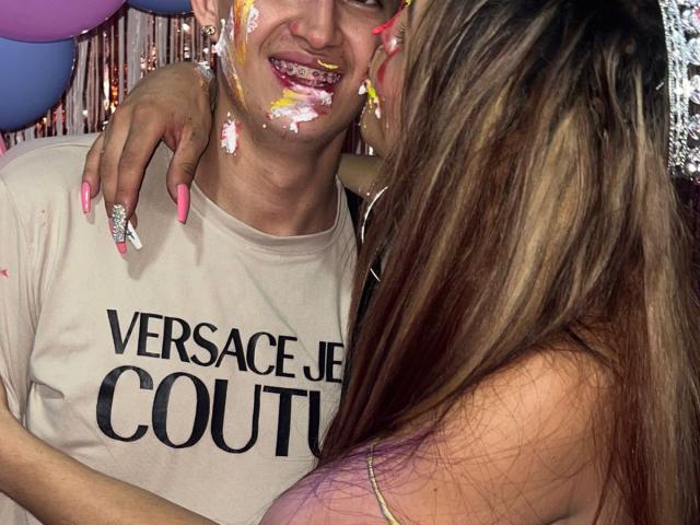 Nahaufnahme eines Fotos von MyaAndJack69 und einer anderen Person. MyaAndJack69 trägt Gesichtsbemalung, ein weißes Hemd mit der Aufschrift "VERSACE JE COUTURE", blaue Nägel, und eine andere Person mit langen, braunen Haaren. sexuelle Spannung, Penis sichtbar.