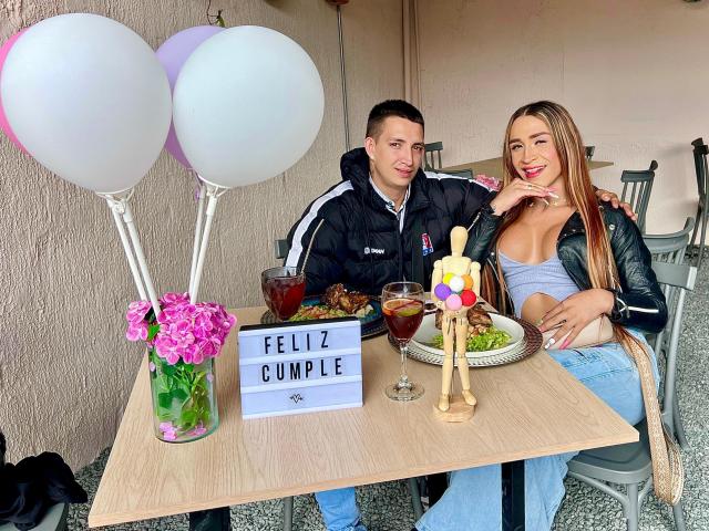 MyaAndJack69 e un uomo sono seduti a un tavolo con due palloncini bianchi, un cartello con la scritta "FELIZ DUMBLE" e dei fiori. MyaAndJack69 indossa un vestito blu e una giacca nera. È visibile il suo inguine..