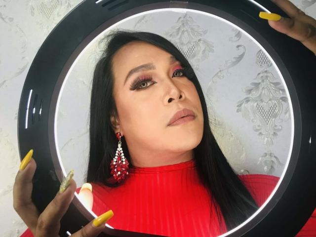 Aziatische vrouw met een groot boezem, lang zwart haar, een rode turtleneck en rode ster-vormige oorbellen, die een ronde ringlamp vasthoudt. Gele nagels. Helder witte achtergrond. Seksuele spanning, verleidelijke blik, direct oogcontact..