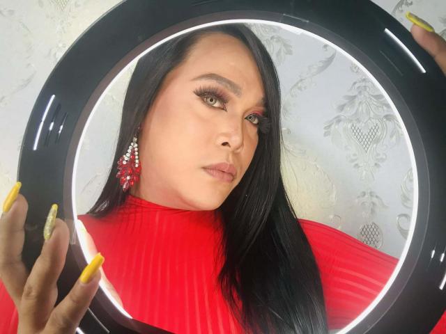 AsianBigBusty with long black hair, wearing a bright red ribbed top, holding a black circular mirror with yellow nails, intense gaze, white patterned background, slight shadow on face.