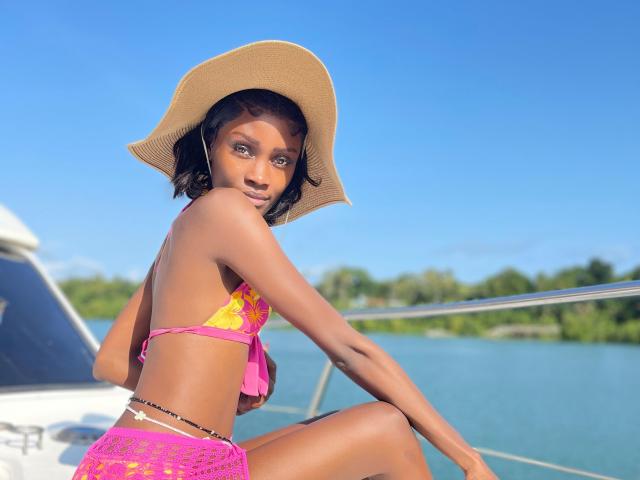 HottieBuddy, avec sa peau bronzée et ses cheveux foncés, porte un chapeau de paille et un bikini floral rose, assise sur un bateau, les jambes écartées. Affichant une pose suggestive et un regard séduisant, avec un fond d'eau bleu vif..