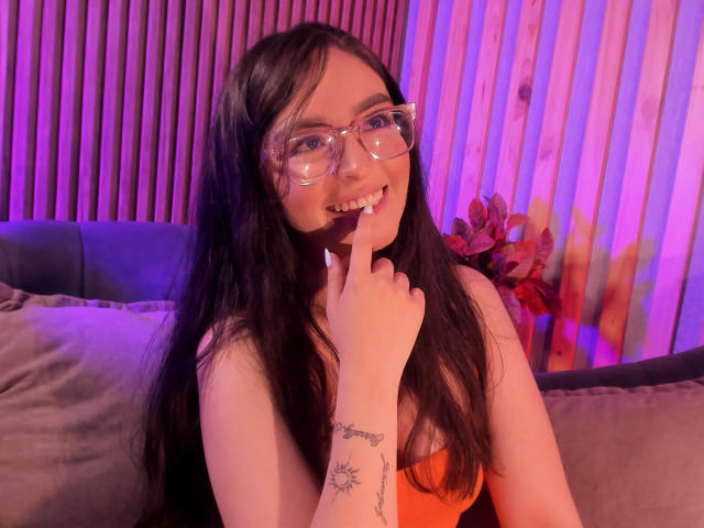 EvaMcAllister, sorridente con un dito sulle labbra, che indossa occhiali, con lunghi capelli neri, una maglietta rossa, un tatuaggio sull'avambraccio, sdraiata su un letto. Sfondo con illuminazione viola e rosa, seducente, sexy, espressione giocosa..