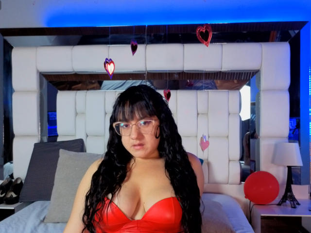 SaraFountain, a curvy woman with long black hair and glasses, wears a red, low-cut top highlighting her large breasts. She's sitting on a bed with white, padded headboard. Blue lighting. Red heart decorations.