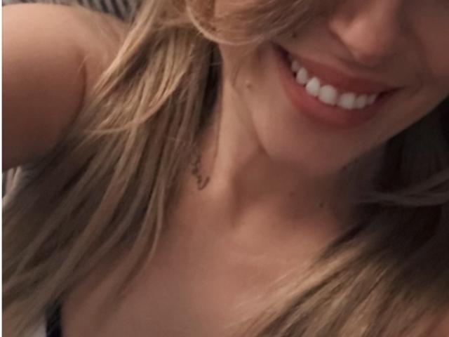 Fotografía de primer plano de XimenaBella, enfocada en su rostro sonriente, mostrando lápiz labial rojo y dientes blancos. El cabello rubio cubre parcialmente su mejilla. La iluminación suave resalta su piel tersa y su expresión segura..