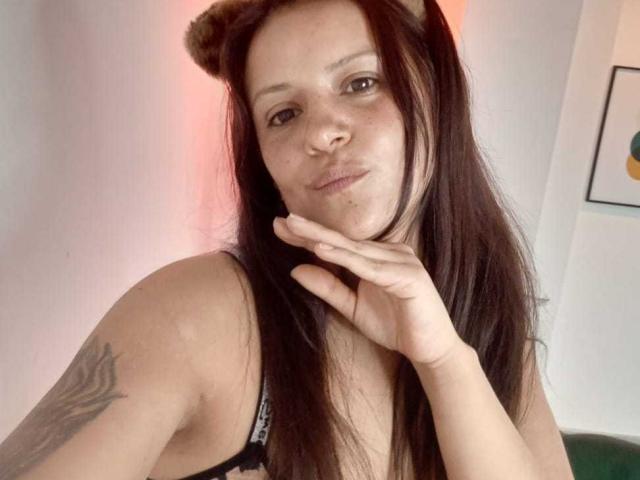 AriannaSeete, mit langen schwarzen Haaren und einem Stirnband mit Bärenohren, posiert verführerisch, die Hand am Kinn, und trägt schwarze Dessous. Tätowierung an der rechten Schulter, leichtes Lächeln, weicher rosa Hintergrund, lockere Selfie-Atmosphäre..