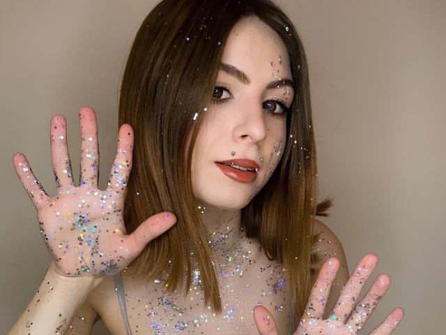 Melita Kiehn, con cabello castaño lacio, piel clara y lápiz labial rojo, levanta ambas manos con los dedos extendidos, cubiertos de brillo de colores. Su expresión es sensual, creando una intensa tensión sexual. Brillo cubre su rostro también..