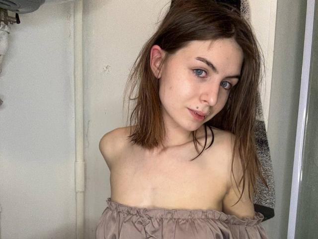 Melita Kiehn hat helle Haut und blaue Augen, mit feuchten, braunen Haaren. Sie trägt ein hellbraunes Oberteil mit einem Träger, das von einer Schulter fällt, und lehnt sich leicht nach vorne, wobei ihre Schlüsselbeine sichtbar sind. Sexuelle Spannung, ein kaum merkliches Lächeln.