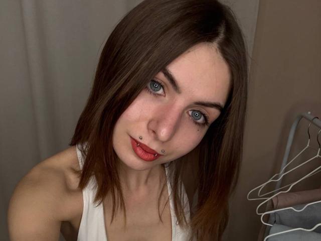 Close-up van MelitaKiehn, een witte vrouw met blauwe ogen, donkerbruin haar en rode lippenstift, die een witte tanktop draagt. Kijkend recht in de camera, met een licht gekanteld hoofd. Heel erg aantrekkelijk..