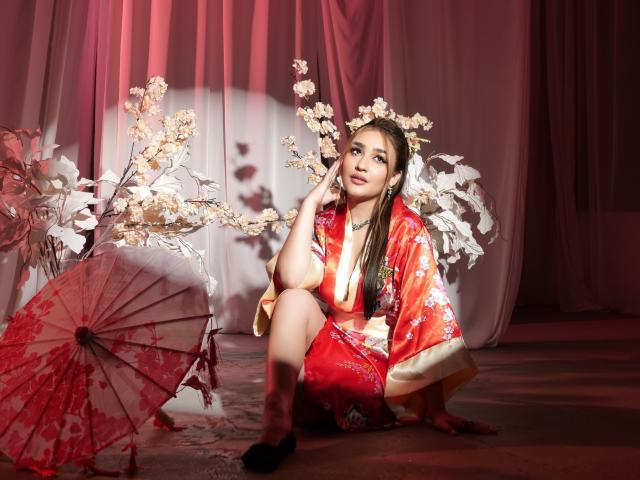 GoldenHear sitzt auf dem Boden in einem roten Kimono, beleuchtet von leuchtend weißen Kirschblüten. Sie hält einen roten Papier-Regenschirm, mit intensiven, sinnlichen Augen und einer anzüglichen Pose..