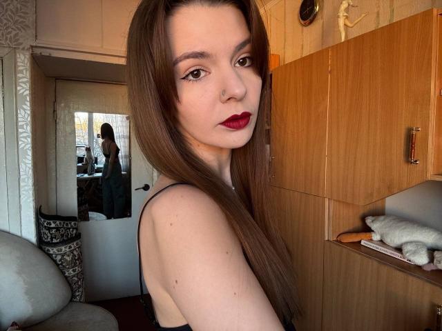 Fotografie von LeidaLala, einer Frau mit heller Haut, langen braunen Haaren, rotem Lippenstift und dunklen Augen, die ein schwarzes Tanktop trägt. Sie blickte über ihre Schulter, mit einem leicht verführerischen Ausdruck. Der Hintergrund zeigt einen Holzzimmer mit einem Bett und einem Spiegel..