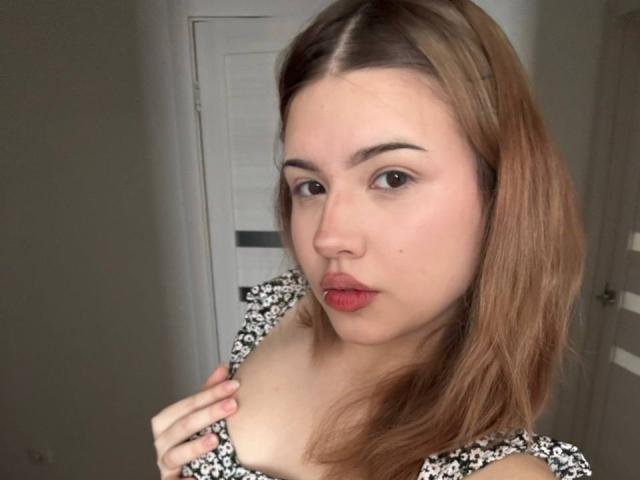 BarrieGromley tiene piel clara y cabello castaño, y lleva un vestido floral en blanco y negro. Ella está sosteniendo su pecho derecho con la mano izquierda, mirando directamente a la cámara con lápiz labial rojo..