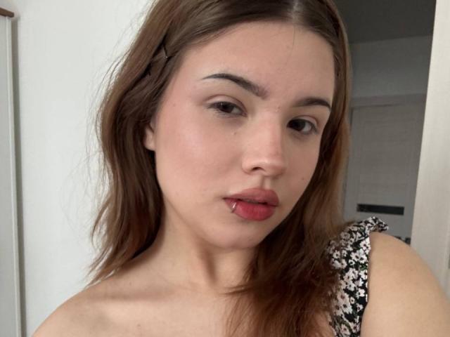 Close-up de BarrieGromley com cabelo castanho longo, pele clara e batom vermelho. Ela olha diretamente para a câmera com os lábios ligeiramente entreabertos. Vestindo um vestido com estampa floral, fundo neutro. Incrivelmente sexy..